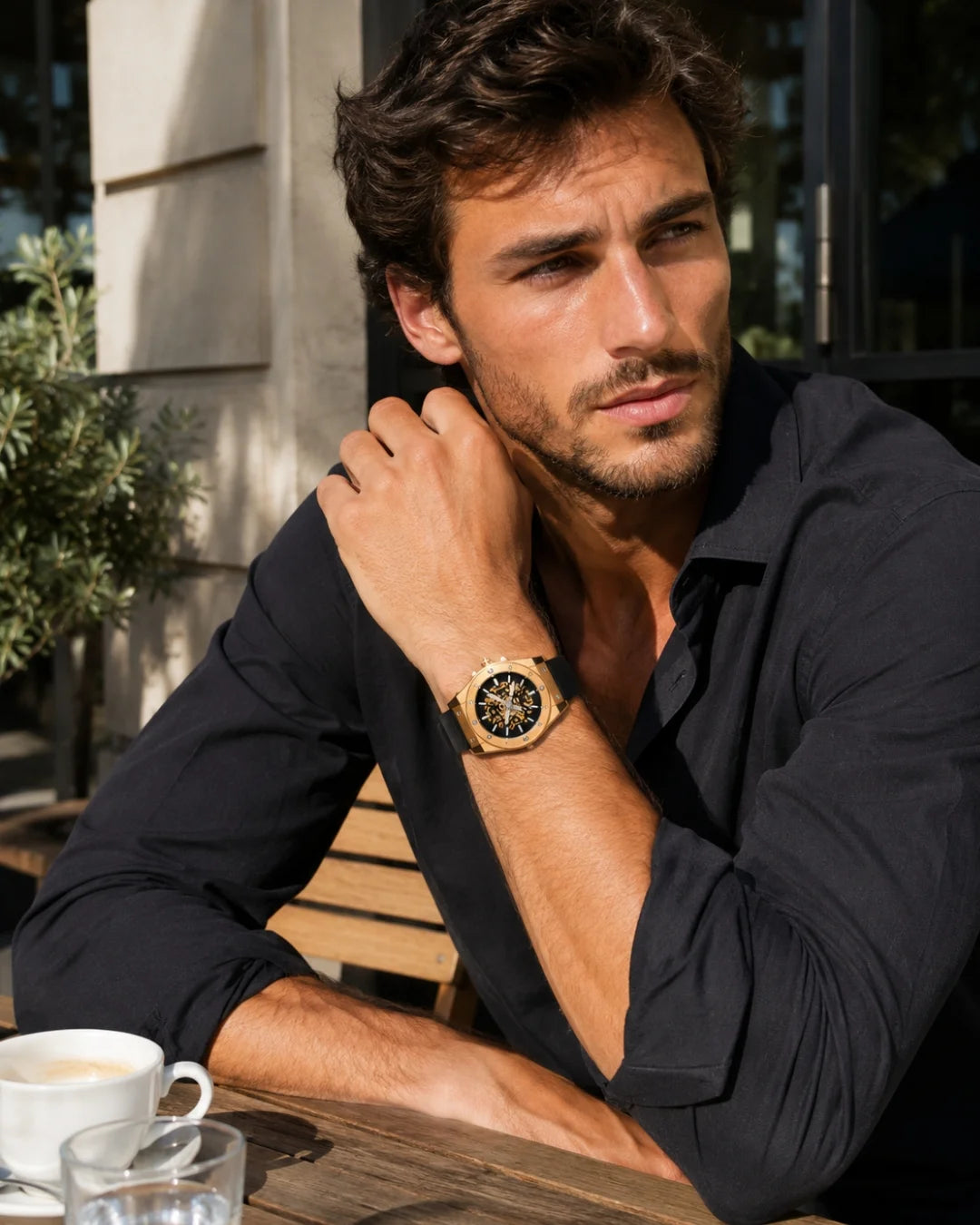 Man in a dark shirt at a cafe table wearing the William Corbier MACH Imperial Gold men's automatic skeleton watch