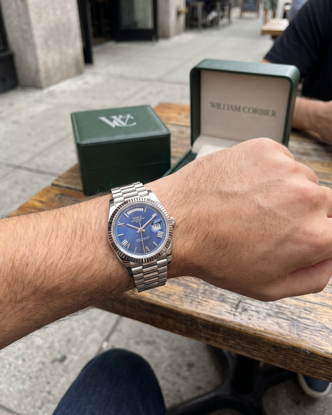 Image of William Corbier NOBLE Dark Azure men's blue dial day-date watch, worn on a wrist beside a green gift box at an outdoor table