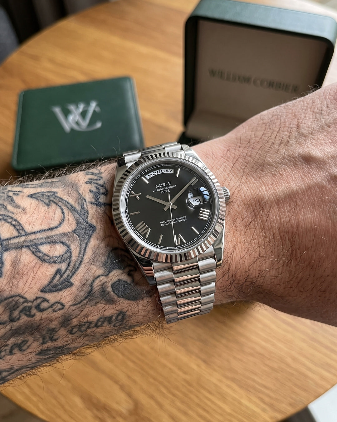 William Corbier NOBLE Obsidian men's black dial day-date watch, worn on a tattooed wrist beside a green gift box