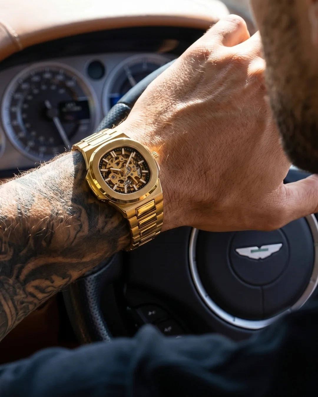 Image of William Corbier PIVOT Imperial Gold men's automatic skeleton watch, worn on a wrist while driving with a tattooed forearm